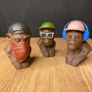 May include: Three small, brown, 3D printed monkey busts with different accessories. The monkey on the left is wearing a black baseball cap and a red bandana. The monkey in the middle is wearing a green hat and white sunglasses. The monkey on the right is wearing a light brown baseball cap and blue headphones.