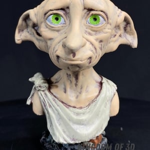 May include: A detailed figurine of Dobby, a house-elf character from the Harry Potter series. The bust features a pale complexion, large green eyes, and a white cloth draped over the shoulders. The base reads "Dobby the Free Elf."