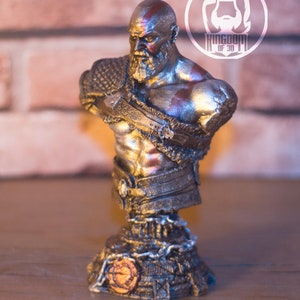 May include: A metallic bust of a bearded warrior with red markings on his head and chest. The bust is on a decorative base with chain details and a circular emblem. The logo "KINGDOM OF 3D" is in the background.