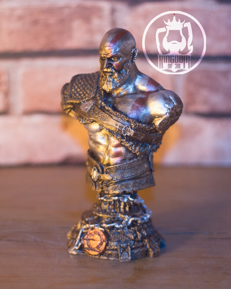 May include: A metallic bust of a bearded warrior with red markings on his head and chest. The bust is on a decorative base with chain details and a circular emblem. The logo "KINGDOM OF 3D" is in the background.