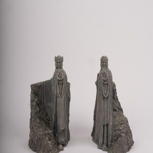 May include: A pair of gray stone-colored bookends featuring sculpted figures of robed individuals standing against jagged rock formations. Each figure is depicted with one arm extended forward, palm open, wearing detailed headgear and long garments.