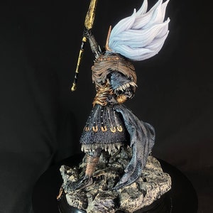 May include: A detailed figurine of a warrior holding a golden spear, wearing a black and gold armor with a flowing white cape. The figurine is standing on a rocky base.