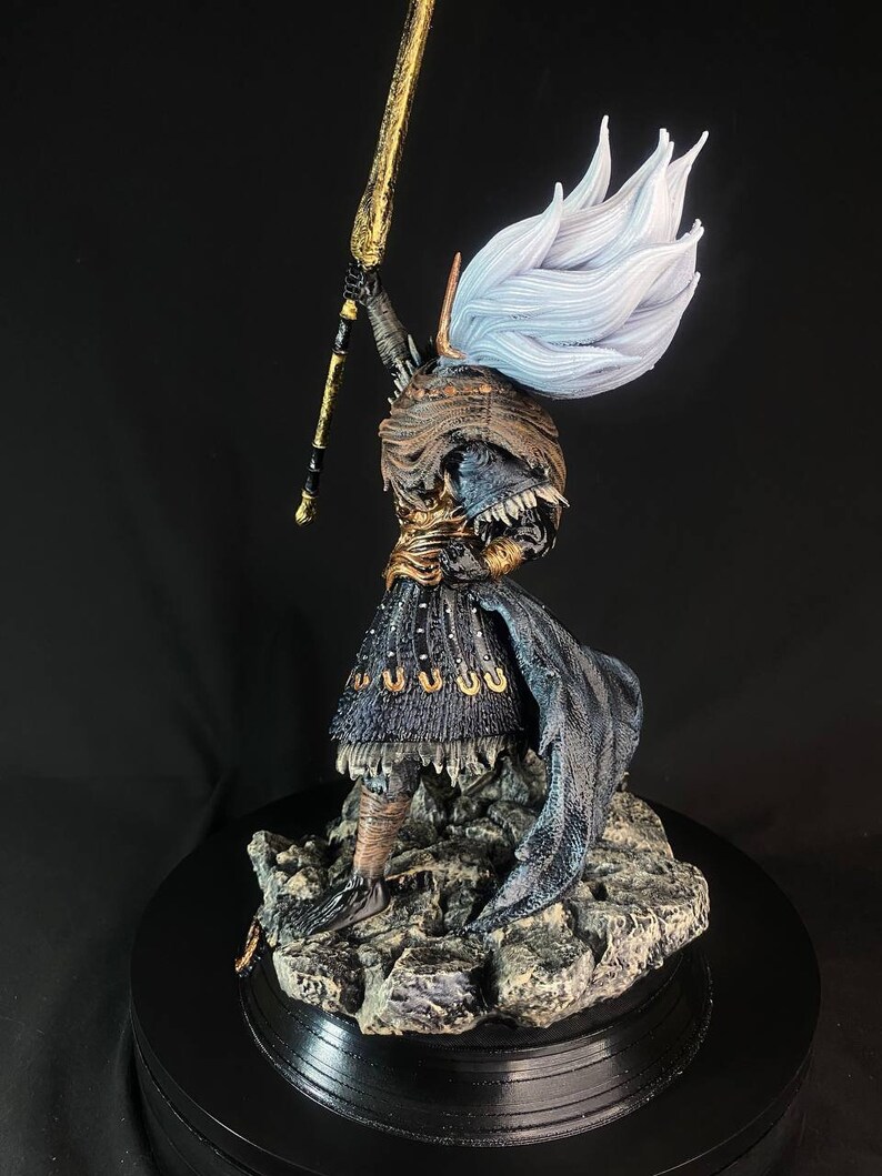May include: A detailed figurine of a warrior holding a golden spear, wearing a black and gold armor with a flowing white cape. The figurine is standing on a rocky base.