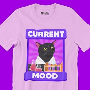 May include: Light pink t-shirt with a graphic featuring a black cat in a lab coat, surrounded by beakers and test tubes. The text "CURRENT MOOD" is displayed above the cat and "MOOD" below. The brand name "NUCLEIC NOODLES" is printed on the collar.