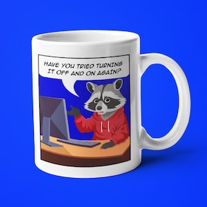 May include: White ceramic mug featuring a cartoon raccoon in a red hoodie sitting at a computer. The raccoon is pointing at the computer screen and a speech bubble above the raccoon says "Have you tried turning it off and on again?"
