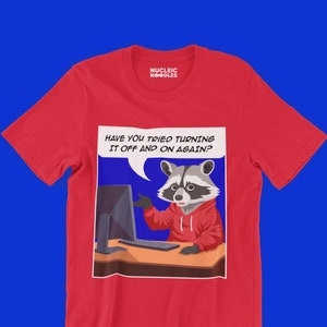 Op de afbeelding: Rood T-shirt met een cartoon wasbeer voor een computer. De wasbeer draagt een rode hoodie en wijst naar het computerscherm. De tekst in de spreekballon zegt "Heb je geprobeerd om het uit en weer aan te zetten?"