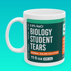 Op de afbeelding: Een witte keramische mok met een groen en oranje etiket met de tekst "0,9% NaCl Biologie Studenten Tranen Normale zoutoplossing 11 fl oz 325 mL".
