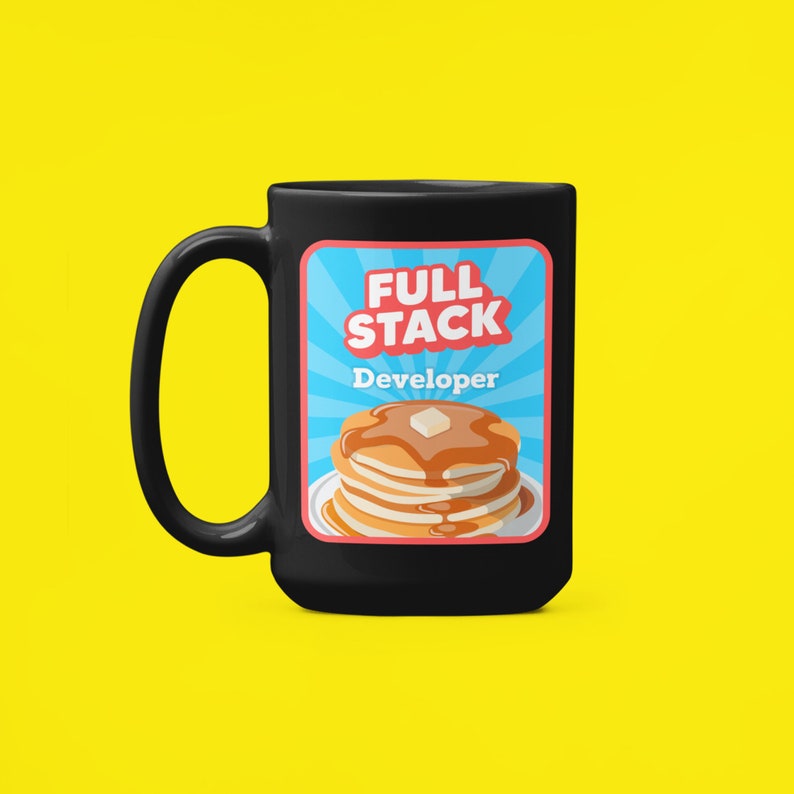 K&ouml;nnte beinhalten: Schwarze Kaffeetasse mit einer wei&szlig;en Grafik eines Stapels Pfannkuchen mit Sirup und einer Butterflocke. Die Grafik enth&auml;lt den Text "Full Stack Developer" in blau und rot.