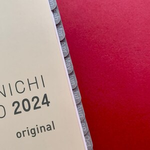 Może przedstawiać: Biały planer z nadrukowanym tekstem "BONICHI CHO 2024 original" na okładce. Planer ma szare zakładki wzdłuż boku oznaczone miesiącami roku: styczeń, luty, marzec, kwiecień, maj, czerwiec, lipiec, sierpień, wrzesień, październik, listopad i grudzień.