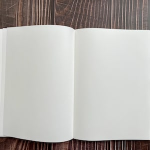 May include: A blank, white notebook with a hard cover, open to a fresh, empty page. The notebook is lying flat on a dark brown wooden surface.