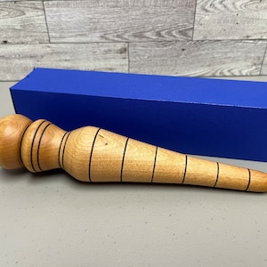 May include: A light-colored wooden massage tool with black accent lines, next to a blue rectangular box. The tool has a rounded head and a tapered handle. The wood grain is visible. The box is a solid, bright blue color.