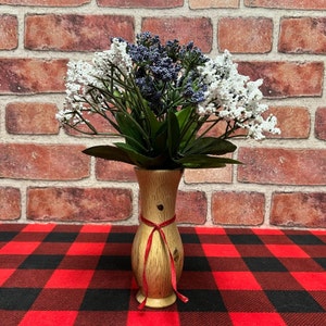 May include: A wooden vase with a red ribbon tied around the neck, holding a bouquet of white and purple flowers. The vase is sitting on a red and black checkered tablecloth.