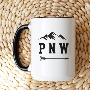 May include: White ceramic mug with a black handle and black rim. The mug features a black silhouette of mountains with the text "PNW" and an arrow pointing to the right.