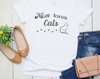 Puede incluir: Camiseta blanca con el texto "Mom loves cats" en fuente cursiva negra. Una silueta negra de un gato está a la derecha del texto. Hay seis huellas de patas negras debajo del texto. La camiseta está colocada sobre una superficie de madera clara con un par de pisos marrones, un par de jeans azules y un bolso marrón claro.