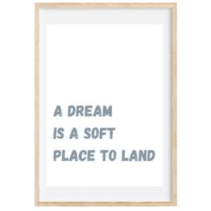 May include: A framed print with the text "A DREAM IS A SOFT PLACE TO LAND" in blue-gray letters. The print is set against a white background and framed in a light wood frame.