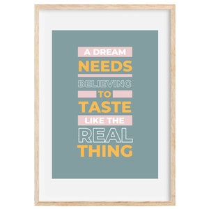 May include: A framed print with a blue background and white text that reads "A dream needs believing to taste like the real thing". The text is in a bold, sans-serif font and is arranged in a vertical column. The words "dream", "needs", "believing", and "thing" are in yellow, while the words "to" and "taste" are in pink.