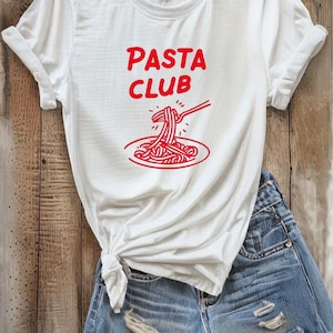 May include: White t-shirt with the words "PASTA CLUB" in red, along with a red graphic of pasta on a plate being eaten with a fork. The shirt is paired with denim shorts and accessories.