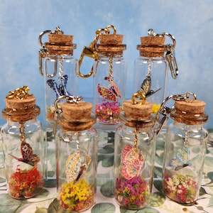 Peut inclure: Sept petites bouteilles en verre avec des bouchons en liège et des porte-clés en métal. Chaque bouteille contient un charme de papillon coloré et des fleurs séchées. Les bouteilles sont disposées en rangée sur une surface verte et brune.