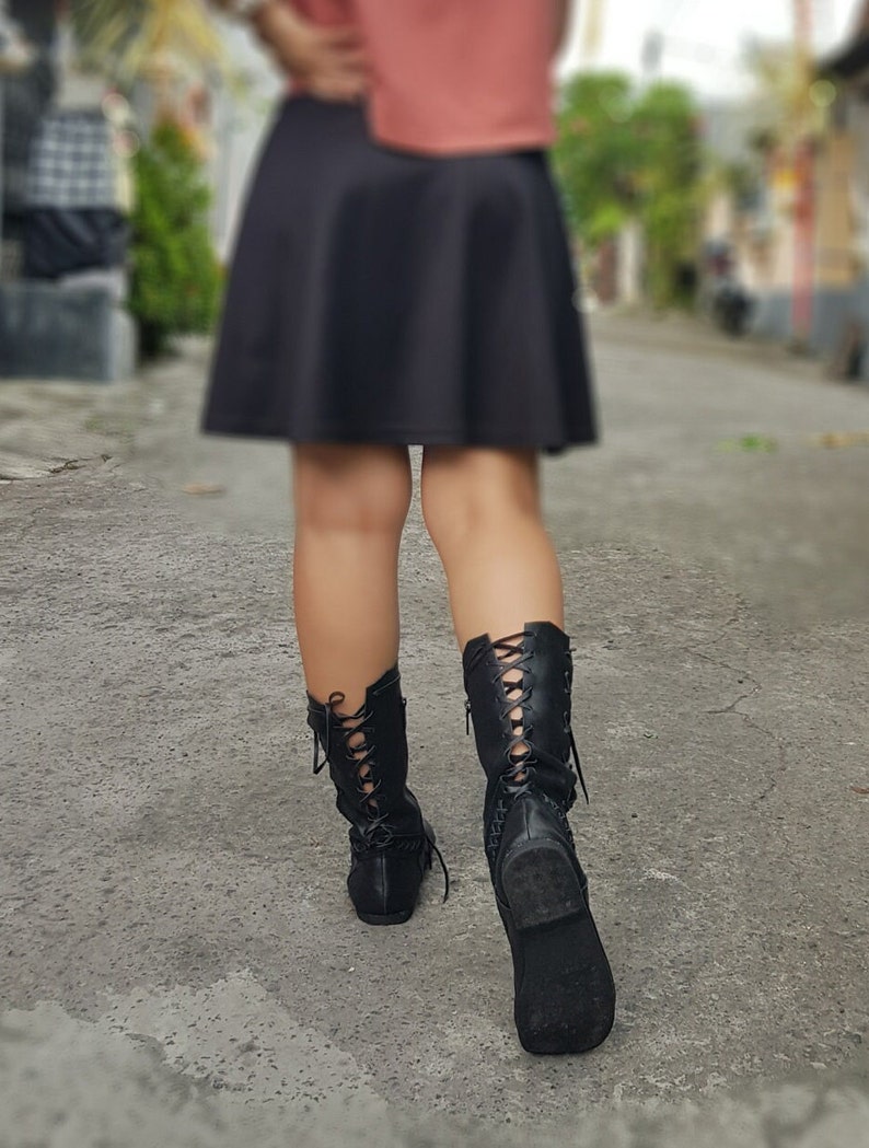 May include: Black lace-up boots with a low heel. The boots have a decorative design on the side and are worn with a black skirt. The boots appear to be made of leather or a similar material.
