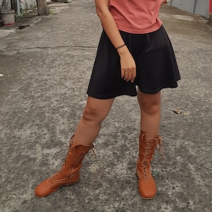 May include: A pair of tan leather boots with intricate lacing and decorative stitching. The boots extend to the mid-calf and have a vintage, Victorian-inspired design. The boots are worn with a black skirt and a peach-colored top.