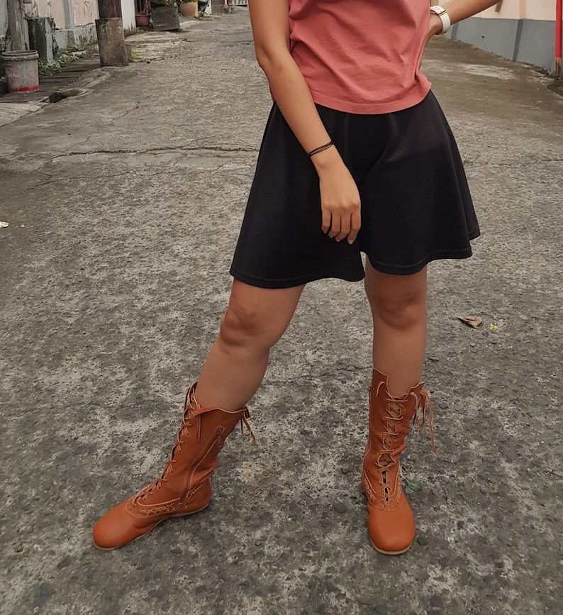 May include: A pair of tan leather boots with intricate lacing and decorative stitching. The boots extend to the mid-calf and have a vintage, Victorian-inspired design. The boots are worn with a black skirt and a peach-colored top.