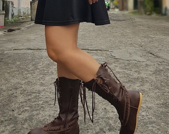 May include: A pair of mid-calf brown leather boots featuring a lace-up front design with decorative side stitching, a side zipper closure, and a flat, light-colored sole. The boots are worn with a black skirt, showcasing the lacing detail.