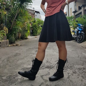 May include: Black knee-high boots with laces and decorative stitching. The boots are worn with a black skirt and a coral-colored top. The image is taken outdoors on a paved road with lush greenery in the background.