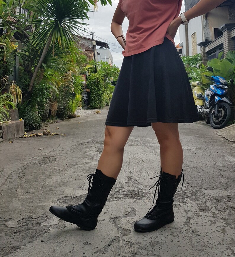 May include: Black knee-high boots with laces and decorative stitching. The boots are worn with a black skirt and a coral-colored top. The image is taken outdoors on a paved road with lush greenery in the background.