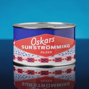 May include: A cylindrical tin of "Oskars Surströmming Fileér" with a blue and red label. The label features the brand name in white, a red diamond shape, and a pattern of fish. The tin is reflected on a glossy surface against a blue background.