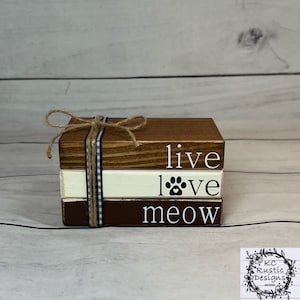 Puede incluir: Un conjunto de tres bloques de madera apilados uno encima del otro. El bloque superior es marrón y dice "live" en letras blancas. El bloque del medio es blanco y dice "love" en letras negras con una huella de pata y un corazón. El bloque inferior es marrón y dice "meow" en letras blancas. Los bloques están atados con cuerda.