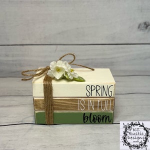 以下が含まれることがあります： 「Spring is in full bloom」と描かれた3つの木製のブロックの積み重ね。ブロックは紐で結ばれ、小さな白い花束で飾られています。