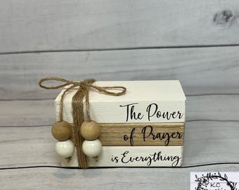 Op de afbeelding: Een decoratieve stapel van drie houten blokken met de tekst "The Power of Prayer is Everything" in zwart schrift. De blokken zijn wit met een natuurlijke houten band en zijn vastgebonden met touw en houten kralen.