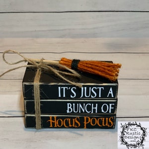 Op de afbeelding: Een stapel van drie zwarte houten blokken met witte en oranje tekst. De bovenste blok leest "It's Just A", de middelste blok leest "Bunch Of" en de onderste blok leest "Hocus Pocus". Een kleine oranje bezem is vastgebonden aan de bovenkant van de stapel met touw.