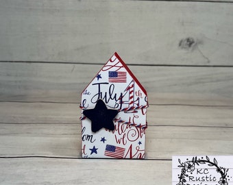 Pode incluir: Uma pequena casa de madeira decorada com cores patrióticas vermelho, branco e azul e uma estrela preta. A casa é decorada com as palavras "the July 4th" e "United States".