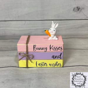 Op de afbeelding: Een driedelige houten blokkenset, geschilderd in roze, paars en geel. De bovenste blok heeft een wit konijnenbeeldje dat een wortel vasthoudt en de tekst "Bunny Kisses". De middelste blok heeft de tekst "and" en de onderste blok heeft de tekst "Easter Wishes".
