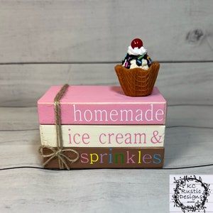 May include: A stack of three wooden blocks with the words "homemade ice cream & sprinkles" painted on them. The top block is pink, the middle block is white, and the bottom block is brown. A small plastic ice cream cone with a cherry on top sits on the top block.
