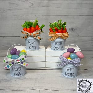 May include: Four white mini buckets with black lettering and pastel ribbon. The buckets are filled with carrots, candy eggs, and the text reads "Bunny Treats", "Fresh Carrots", "Easter Eggs", and "Happy Easter".
