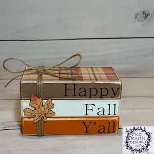 Könnte beinhalten: Ein Stapel aus drei Holzblöcken mit den Worten "Happy Fall Y'all". Der obere Block hat ein Karomuster, der mittlere ist weiß und der untere ist orange. Eine Schnur-Schleife und ein Blatt schmücken die Blöcke.