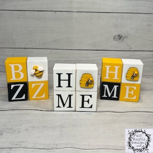 May include: Wooden block letters spelling "HOME" with a bee and honeycomb design on each block. The blocks are painted white with black letters and yellow accents.