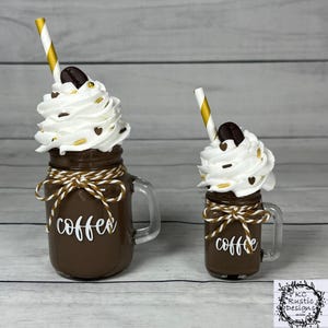 Peut inclure: Deux tasses à café décoratives avec de la crème fouettée, des grains de café et des pailles dorées. Les tasses sont marron avec le mot "coffee" en écriture blanche. Chaque tasse est attachée avec de la ficelle marron et dorée.
