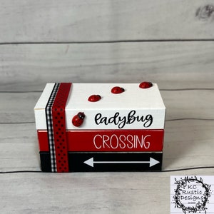 Puede incluir: Una caja de madera de tres niveles con un esquema de color rojo, blanco y negro. El nivel superior es blanco con las palabras "ladybug crossing" en negro. El nivel intermedio es rojo con lunares blancos. El nivel inferior es negro con flechas blancas. Hay cuatro mariquitas rojas en el nivel superior.