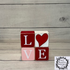 Puede incluir: Bloques de madera que deletrean la palabra "LOVE" con un corazón rojo en el centro. Los bloques están pintados de rojo, blanco y rosa.