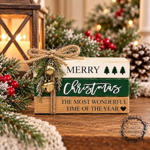 May include: A stack of three rectangular wooden blocks tied with twine and decorated with faux greenery and two gold jingle bells. The top block is white with MERRY and three tree icons. The middle is green with Christmas. The bottom is natural wood with THE MOST WONDERFUL TIME OF THE YEAR and a heart icon.