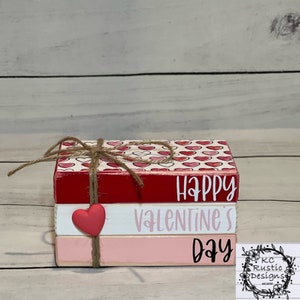 May include: A stack of three wooden blocks with the words "Happy Valentine's Day" painted on them. The top block has a red heart pattern, the middle block is white, and the bottom block is pink. A small red heart is attached to the middle block.