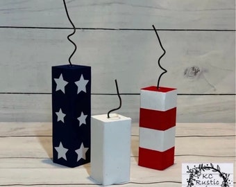 May include: Three wooden blocks painted in red, white and blue, resembling fireworks. Each block has a black wire attached to the top, resembling a fuse.