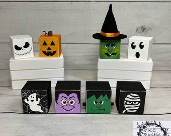 May include: A selection of Halloween-themed wooden blocks. The blocks are decorated with spooky characters such as a Jack Skellington face, a carved pumpkin, a witch, a ghost, a vampire, a Frankenstein monster, and a mummy. The blocks are painted and sit on white platforms.