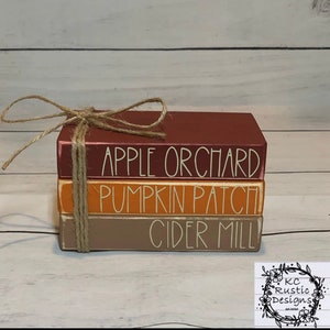 May include: A stack of three wooden blocks painted in fall colors with the words "Apple Orchard", "Pumpkin Patch", and "Cider Mill" in white paint. The blocks are tied together with twine.