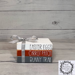May include: A stack of three wooden blocks with the words "Easter Eggs", "Carrot Patch", and "Bunny Trail" painted on them. The blocks are painted in white, orange, and gray. The blocks are tied together with a white and gray checkered ribbon.