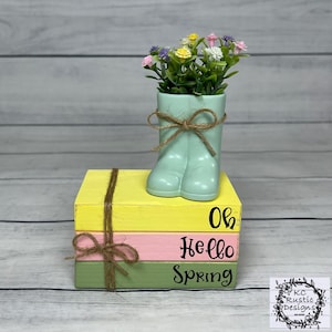 Op de afbeelding: Een decoratief lentestuk. Een lichtgroene laarzenvormige vaas met kunstbloemen staat op drie gestapelde, geverfde houten blokken. De blokken zijn geel, roze en groen, met de tekst "Oh Hello Spring".