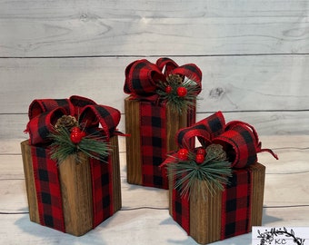 May include: Three wooden boxes wrapped in red and black buffalo plaid ribbon with a bow and greenery. The boxes are decorated with pine cones and red berries.